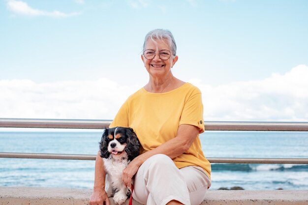 Old senior woman in yellow jersey sitting close to the beach with her cavalier king charles dog