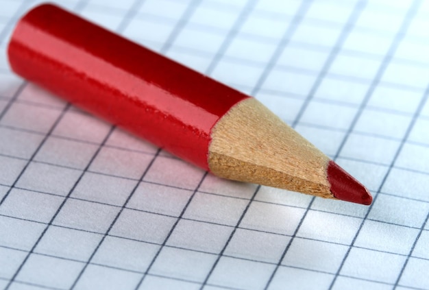 An old small used sharpened red wooden slate pencil lies on a checkered paper sheet, close-up