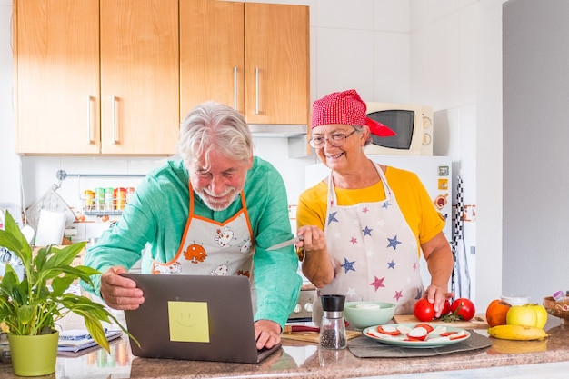 Old spouses enjoy vegetable salad preparation together in kitchen. Caring grey-haired husband feeding loving wife, romantic date, happy marriage, healthy eat concept