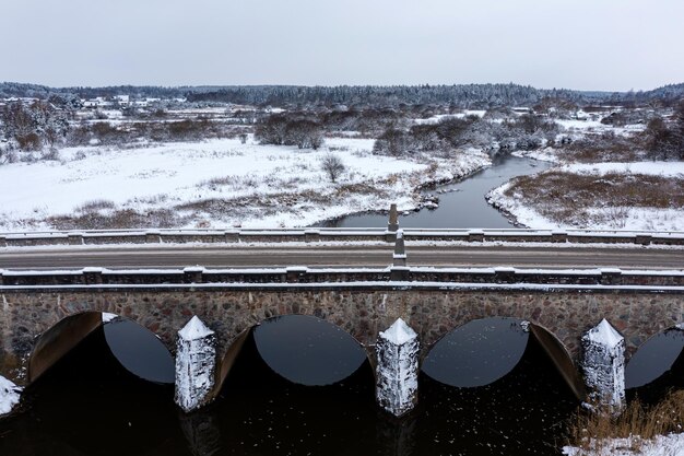 雪の降る冬の日にアババ川に架かるアーチのある古い石造りの橋、カンダヴァ、ラトビア