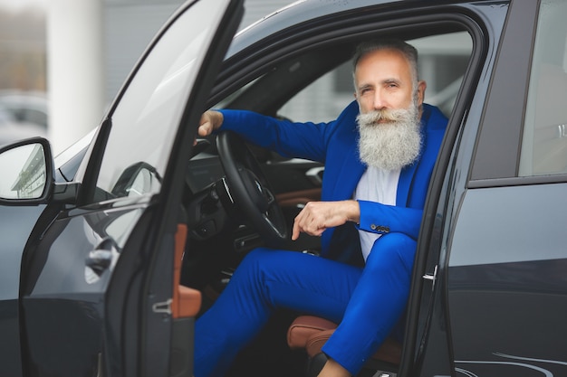 高級自動車の近くに立っているお洒落な老人。車の近くの屋外でスーツを着た成熟した金持ちの男性。