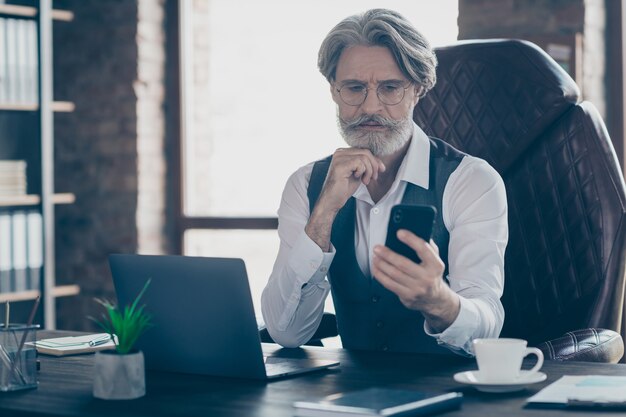 Old thoughtful businessman sit desk using smartphone in office