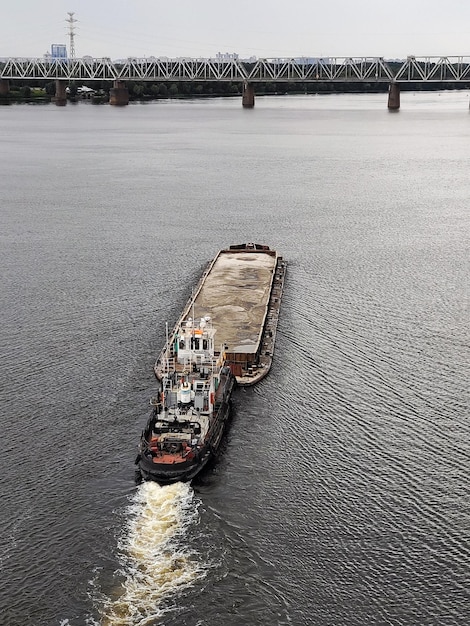 An old tugboat pushes a barge with sand in front of it and\
floats down the river there is an iron bridge ahead