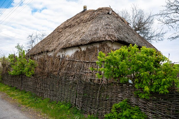 ウクライナの田舎風の古いヴィンテージの田舎の家。ガマの屋根の家。葦の屋根。小屋の近くの枝編み細工品の柵（編み枝細工の柵）。