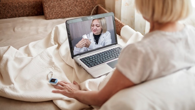 Vecchia donna a letto guardando lo schermo del laptop e consultando un medico online a casa, servizi di telemedicina durante il blocco, videochiamata distante, moderna applicazione sanitaria tecnologica