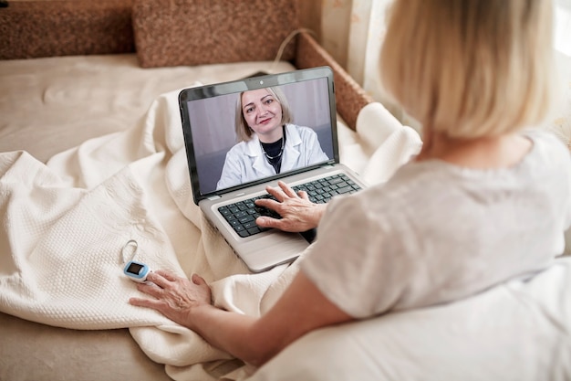 Foto vecchia donna a letto guardando lo schermo del laptop e consultando un medico online a casa, servizi di telemedicina durante il blocco, videochiamata distante, moderna applicazione sanitaria tecnologica