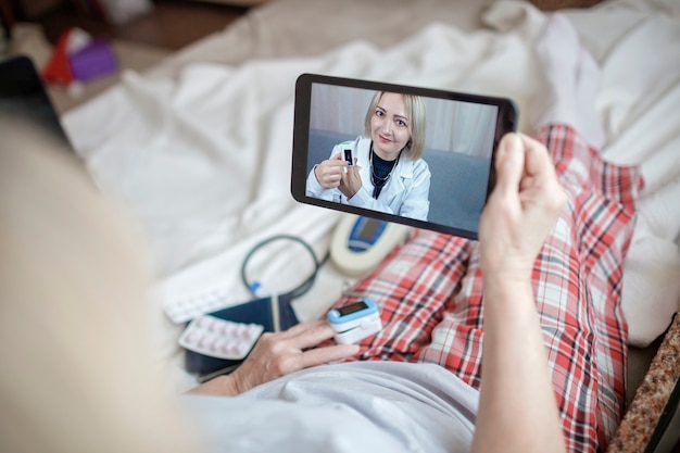 Old woman in bed looking at screen of laptop and consulting with a doctor online at home, telehealth