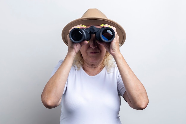 Old woman in a hat looking through binoculars on a light background.