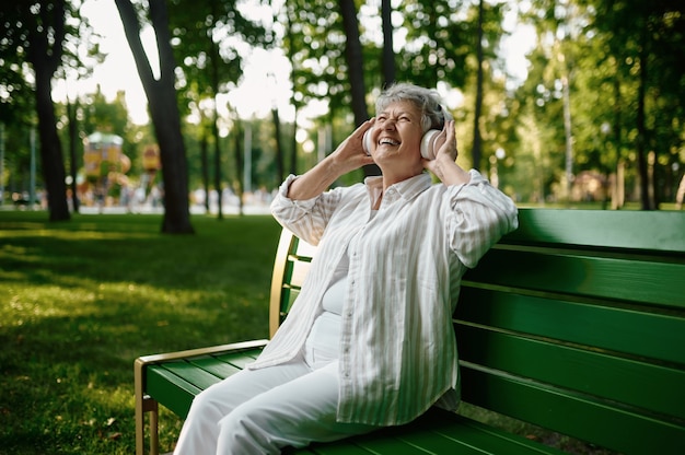 Old woman in headphones listens to music on the bench in summer park. Aged people lifestyle. Pretty grandmother having fun outdoors, an elderly female person outdoors
