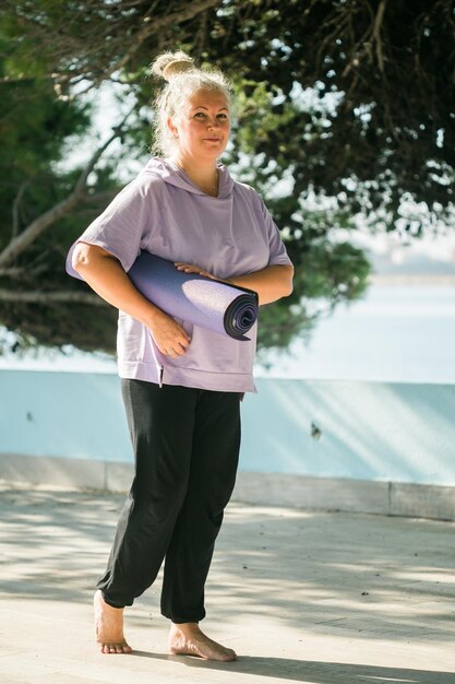 Old woman holding yoga mat and preparing to practice yoga or meditation outdoors on sea beach Happy mature woman with dreadlocks exercising on seashore Meditation or yoga and relaxation concept