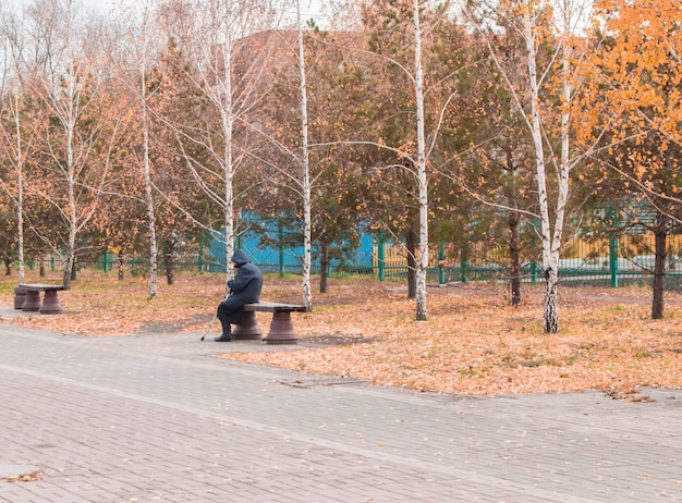 Старуха с палкой сидит на скамейке в парке.