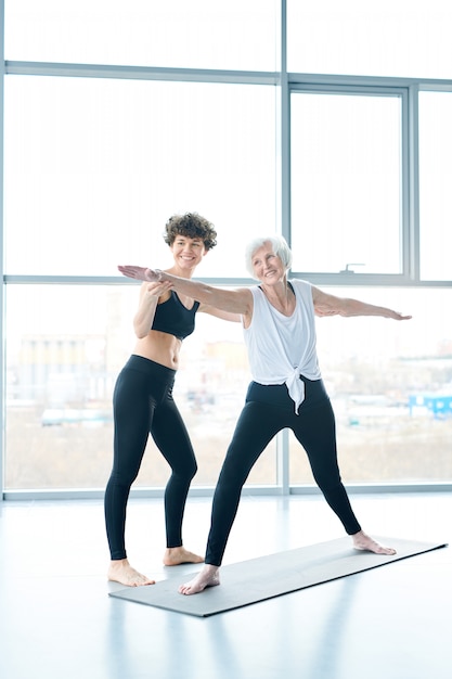 Old woman on mat doing yoga next to a large window