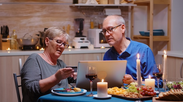 Old woman paying online the holiday using laptop and card during romantic dinner. Old people sitting at the table, browsing, using the technology, internet, celebrating their anniversary.