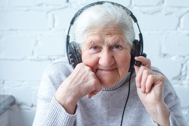 An old woman sits at a table in front of a laptop with headphones.