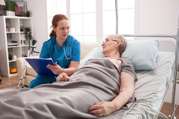 Photo old woman sitting in bed in nursing home talking with female doctor with clipboard.