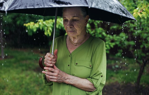 An old woman stands on the street under the rain with a black umbrella