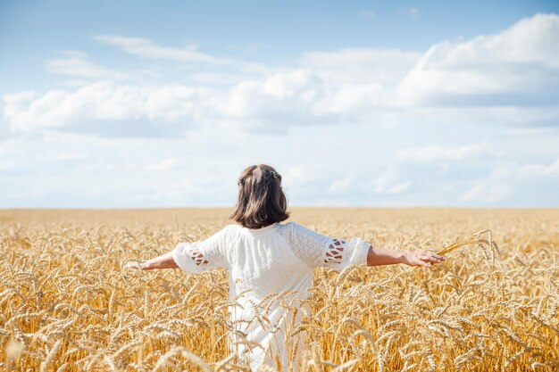 Old woman stands with her back with her arms spread out to side in field with golden ears of cereal crops xDxA