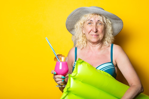 Old woman in swimsuit with cocktail holds air mattress on yellow surface