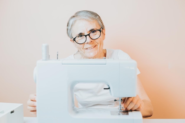 ミシンに取り組んでいる年配の女性の仕立て屋は、縫いながらカメラに幸せそうに微笑んでいます 新しい服の持続可能性のために古い生地を再利用します 高齢者の家庭での新しい趣味 高齢者の精神活動と仕事