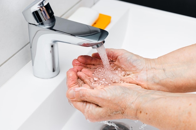 Old woman washing hands under streaming water from faucet without soap in bathroom hygiene concept