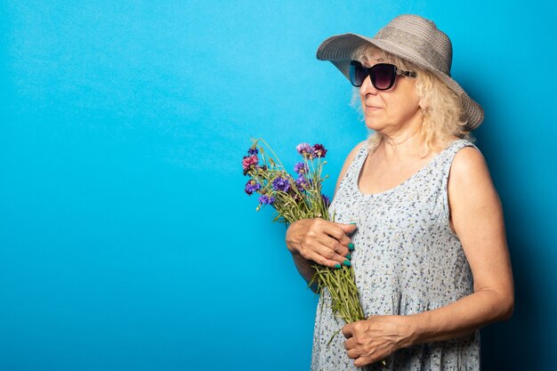 Old woman in a wide-brimmed hat and dress holds a bouquet of flowers and looks to the side on a blue wall.