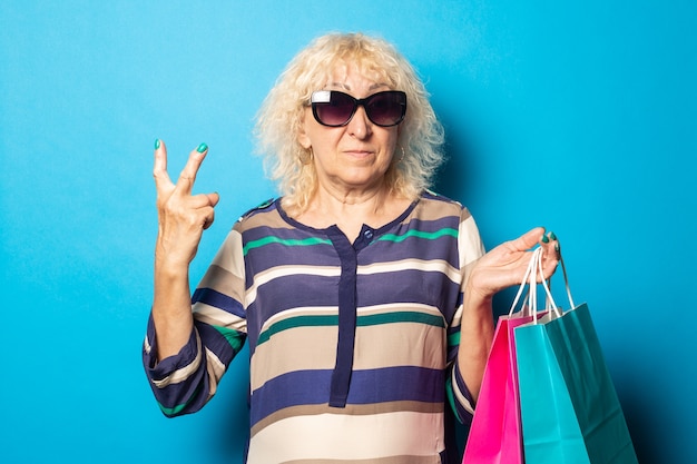 Old woman with glasses holds shopping bags and shows friendly, welcome gesture on blue wall.