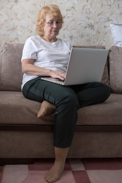 Old woman with glasses sits on the sofa and works on laptop at home, vertical orientation. Remote work