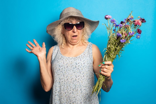 Old woman with a surprised face in a wide-brimmed hat and dress holding a bouquet of flowers on a blue surface