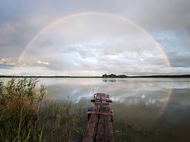 の曇り空の風景の美しい虹の背景にある湖の古い木製の橋