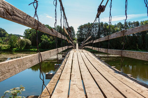 Старый деревянный подвесной мост на реке в солнечный летний день. Природный ландшафт. Перспектива