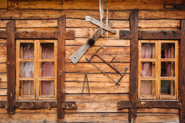 An old wooden wall of a house with windows in the village and tools hanging