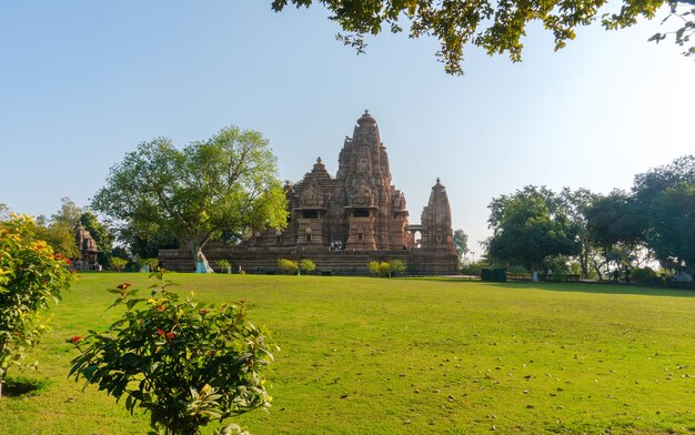 Old and younger Hindu temple, built by Chandela Rajputs, at Western site in India's Khajuraho framed by trees. White grey for the younger and older beige structure against blue skies over green grass.