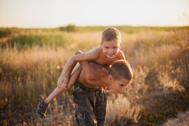 The older brother carries his laughing younger brother on his back in the summer field at sunset