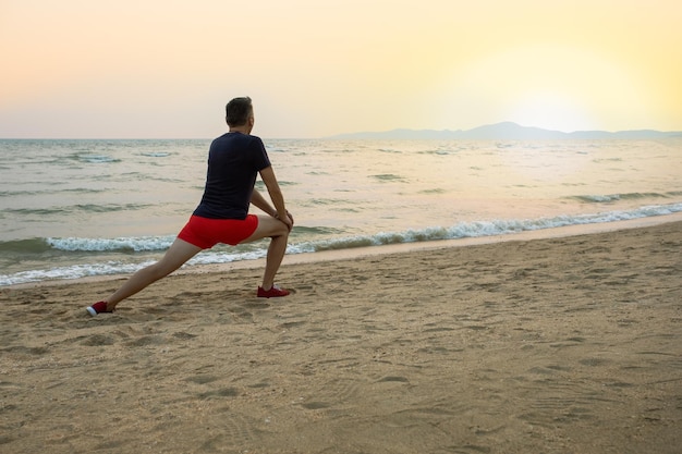 Older man doing stretching legs exercises on the beach at sunrise or sunset