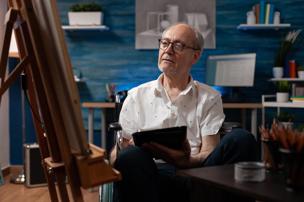Older man sitting in wheelchair holding digital tablet computer looking at vase sketch drawing on easel. Retired artist living with disability using modern technology looking for inspiration.