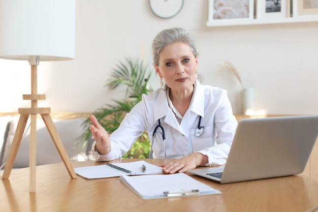 Older woman doctor therapist wearing headset video call talking\
to web camera consulting virtual patient online by video conference\
call chat.