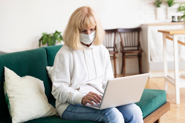 Donna anziana a casa che lavora al computer portatile con mascherina medica