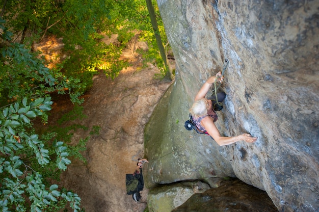 Older woman rock climber is climbing with carbines and rope on a rocky wall of big rock. 