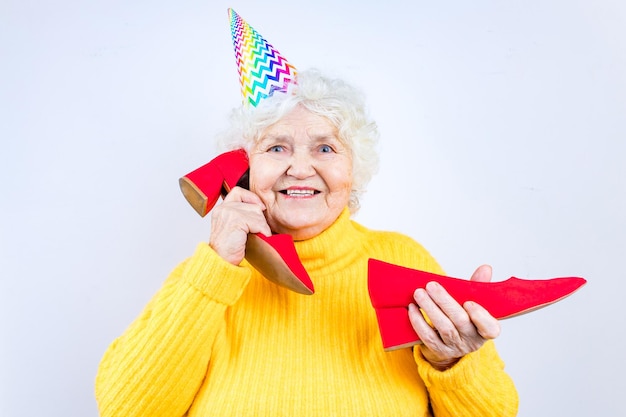 Older woman with a gift wear yellow sweater and horn cap on a white background holding red high heels shoes