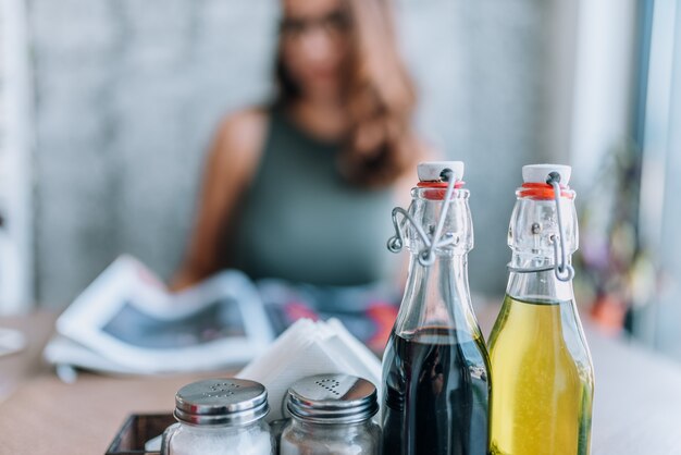 Olie, balsamico azijn, zout en peper op tafel. vage vrouw die een tijdschrift op de achtergrond leest.