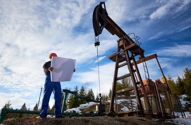 Foto oliearbeider die zich in het olieveld naast een pompkrik bevindt met een groot papier