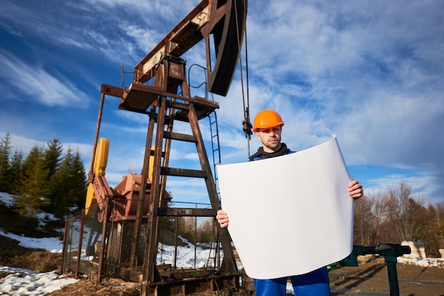 Oliearbeider die zich in het olieveld naast een pompkrik bevindt met een groot papier