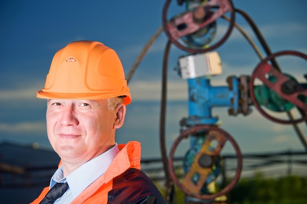 Oliearbeider in oranje uniform en helm op de achtergrond van de kleppen, leidingen en avondrood.