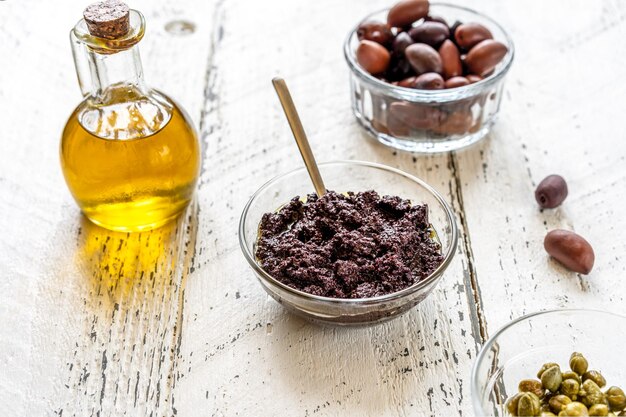 Olijfpastei in kom en basisingrediënten olijfolie, olijven en kappertjes op witte houten ondergrond