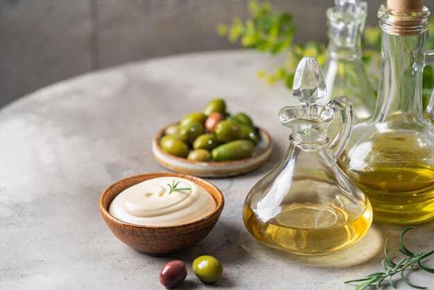 Photo olive oil in a glass bottle containers mayonnaise sauce and harvest olives with herbs on gray background