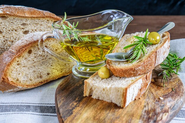 Foto olio d'oliva nella ciotola di vetro trasparente sul tagliere di legno. pane integrale affettato, olive e rosmarino. concetto di cibo mediterraneo sano.