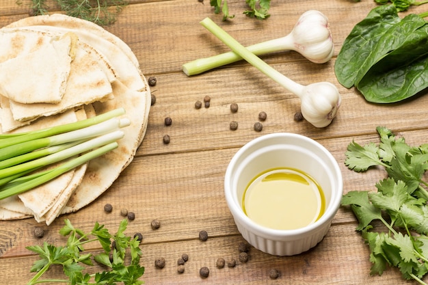 Olive oil in white bowl. Green onions on pita bread. Garlic, greens and sweet peas on table. Vegetarian snack. Light wooden background. Top view.