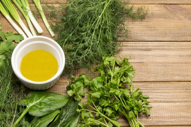 Olive oil in white bowl Green onions spinach parsley and dill on table Copy space Light wooden background Top view