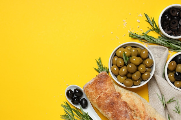 Olives in bowls and baguette on yellow background space for text