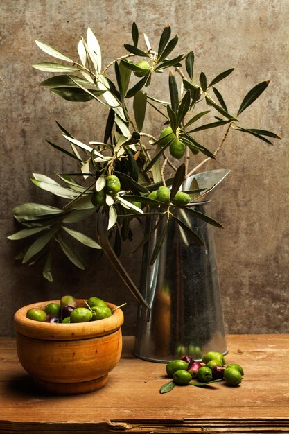 Olives in a wooden bowl and a metal jug with olive branchs with leaves on a wooden table